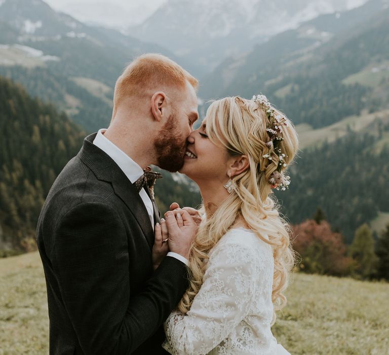 Boho bride in an ethereal wedding dress with lace bodice, half up half down wedding hair and flower crown kissing her groom in a check suit