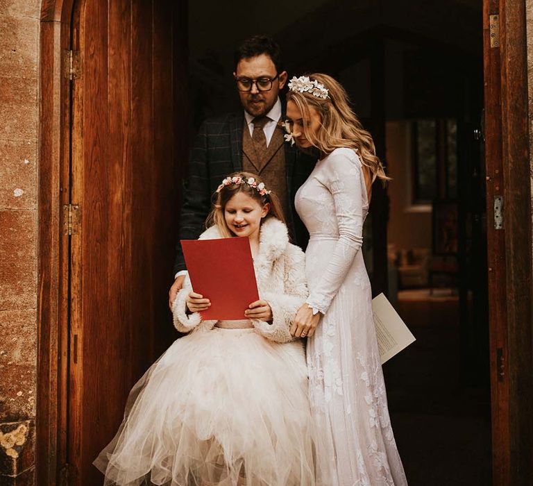 Bride, groom and their daughter in a tutu and faux fur coat holding a red envelope 