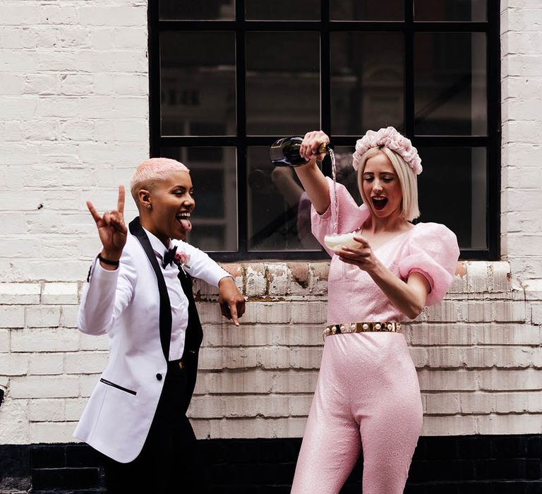 Bride in a shimmering pink jumpsuit with peg leg trousers and puff sleeves during champagne for her bride in a white tuxedo 