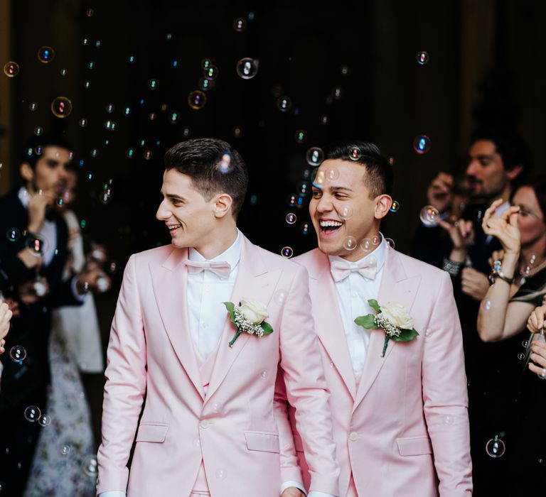 A gay couple leave their wedding ceremony hand in hand and laughing. They wear matching pink suits. Photography by Michael Maurer.