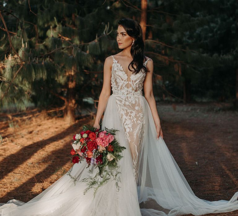 Bride wearing a lace embroidered wedding dress with tulle overskirt, carrying a bouquet of red and pink peonies and roses