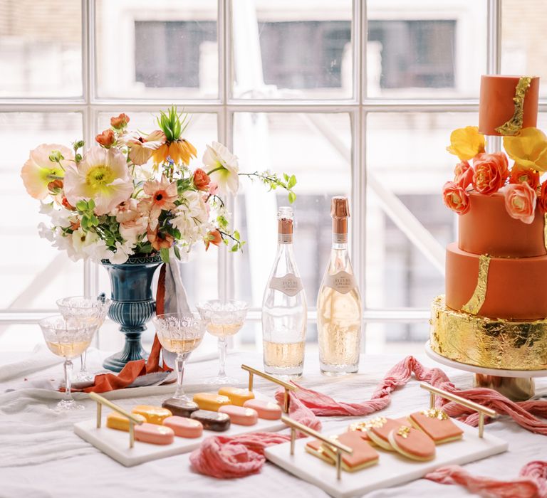 Orange wedding cake on white table cloth next to floral display and dessert favours in pastel colours