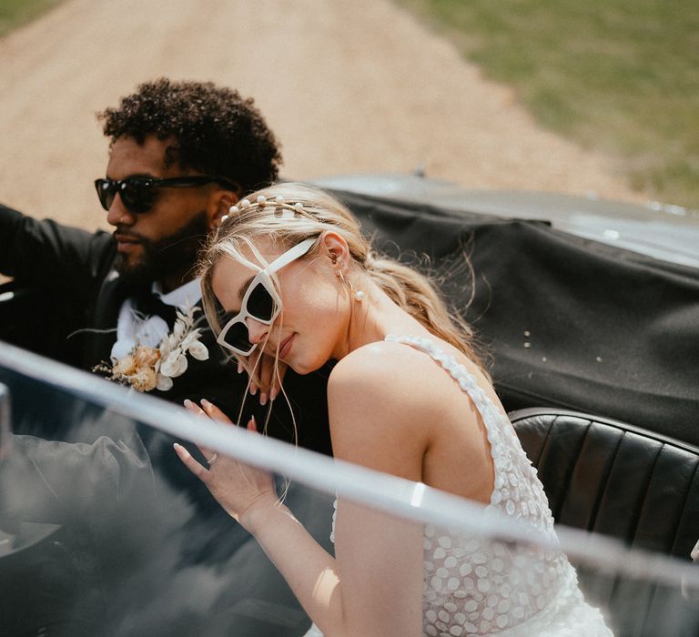 Bride in an appliqué wedding dress, peal headband and white sunglasses resting her head on her grooms shoulder as they sit in their vintage wedding car 