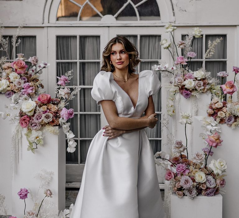 Stylish bride in a plunging neckline wedding dress with puff sleeves standing in front of the wedding ceremony floral arrangements with pink and white orchids and roses 