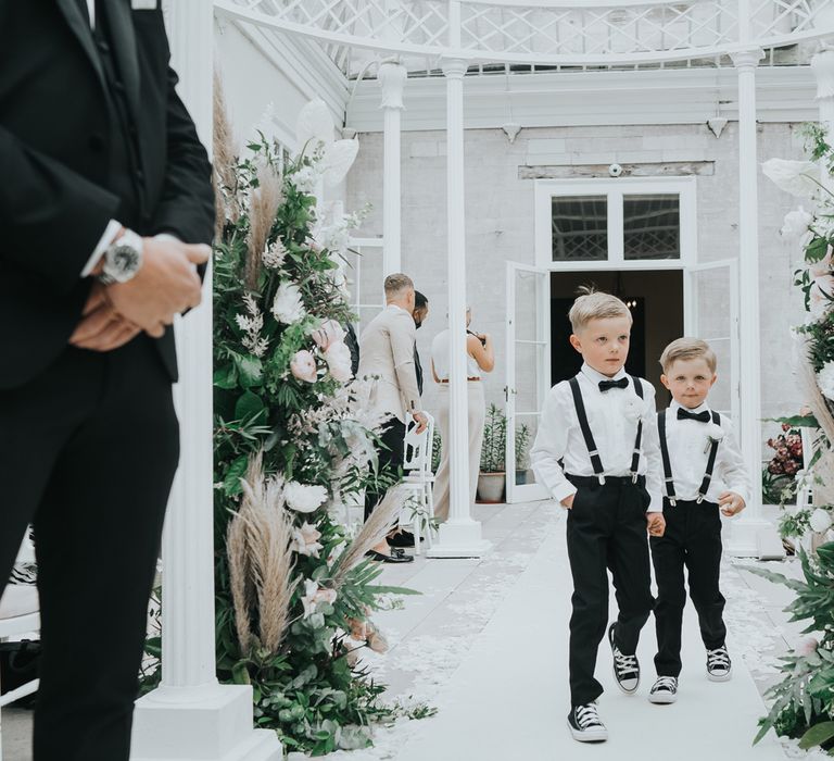 Two young boys in white shirts, braces, bow ties and black and white converse walk down white carpeted aisle hand in hand in the glass conservatory during wedding ceremony at Came House Dorset