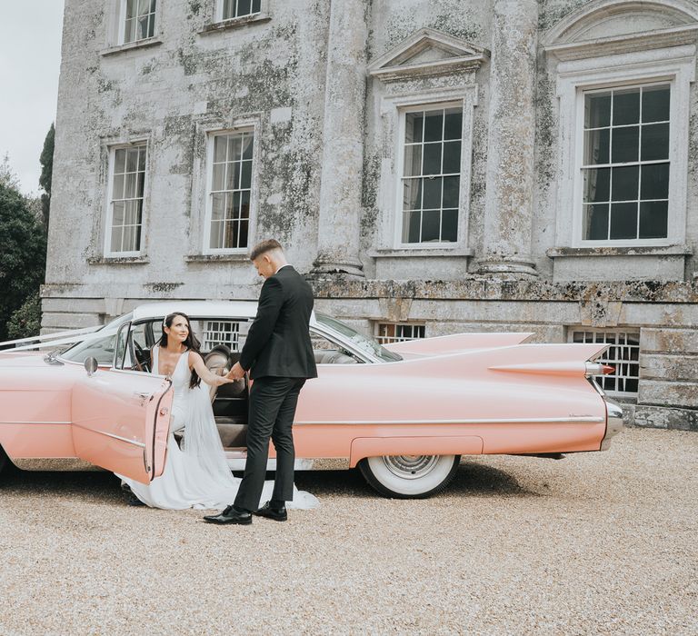 Groom in black tuxedo helps bride in white Pronovias wedding dress out of pink Cadillac outside the front of Came House Dorset 