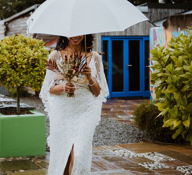 Bride walks down path whilst holding white umbrella and dried floral bouquet 