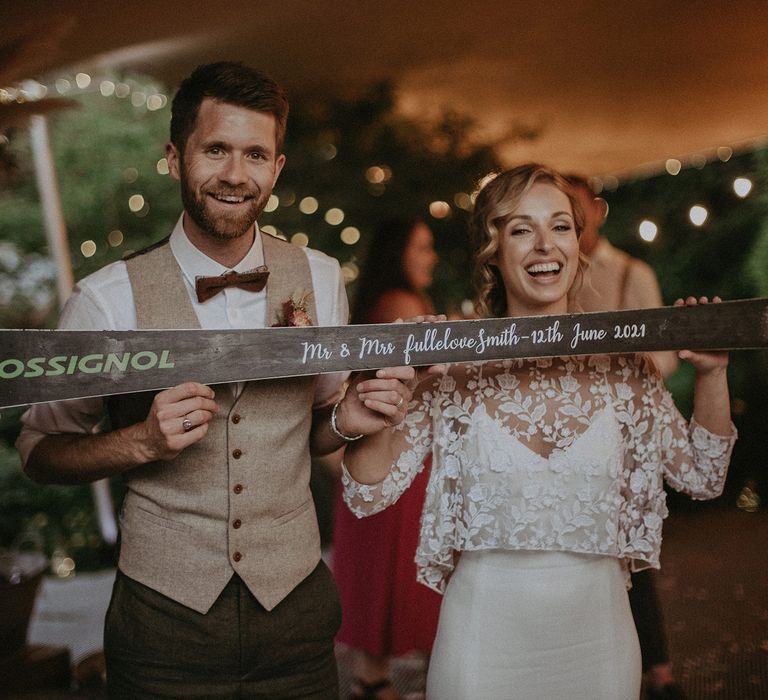 Bride in white Rime Arodaky wedding dress with mesh floral embroidered top and groom in white shirt and light brown waistcoat and bowtie both smile whilst holding up ski shot with writing on the bottom