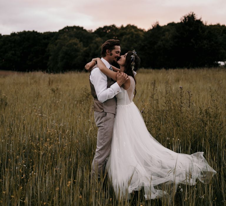 Bride & groom embrace outdoors in green fields as the sun begins to set turning the sky pink