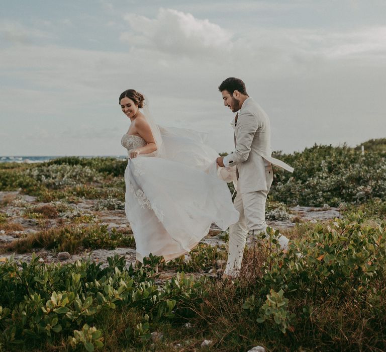 Bride & groom walk along greenery as groom holds her wedding gown and smiles 