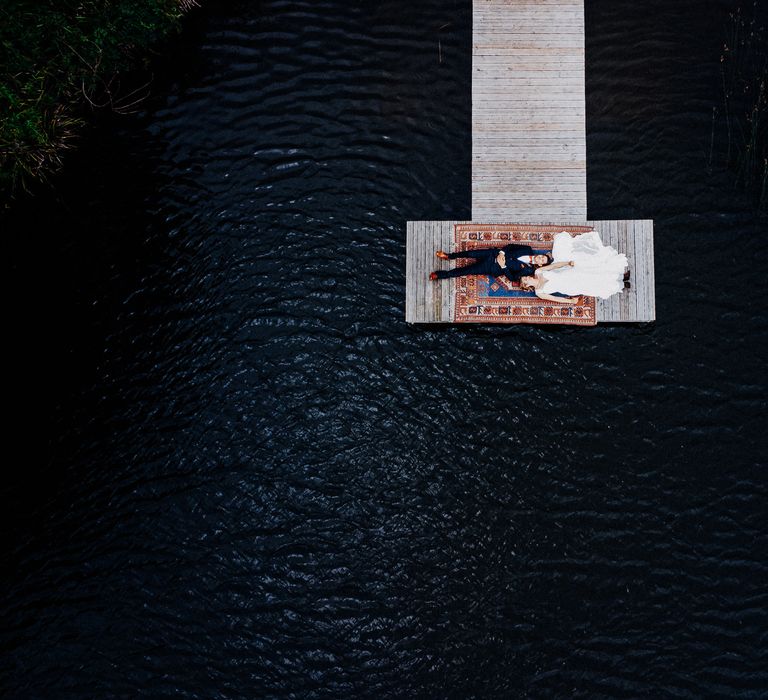 Drone image of couple lay with one another on jetty on a multicoloured rug before wedding ceremony