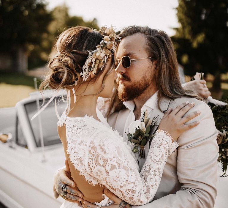 Bride in a long sleeve wedding dress and pinned bridal up do embracing her groom in a beige suit by the side of their white wedding car 