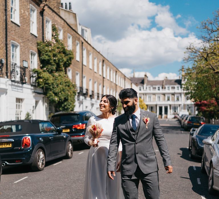 Bride & groom walk through Chelsea as the sun shines and bride holds small dried flower bouquet