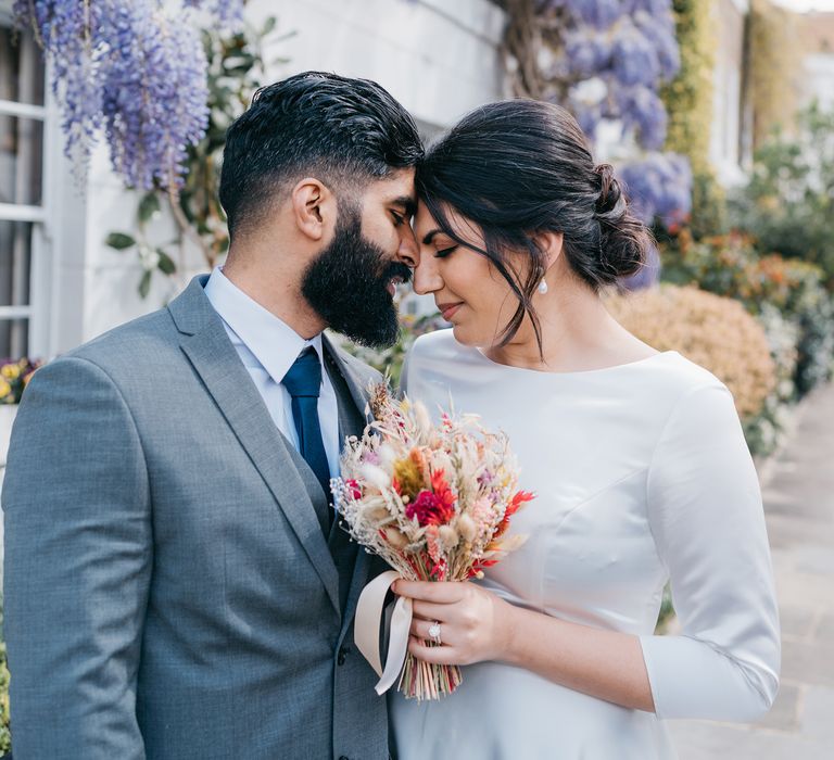 Bride & groom lean in to one another on their wedding day as they stand in front of hanging purple wisteria tree in Chelsea