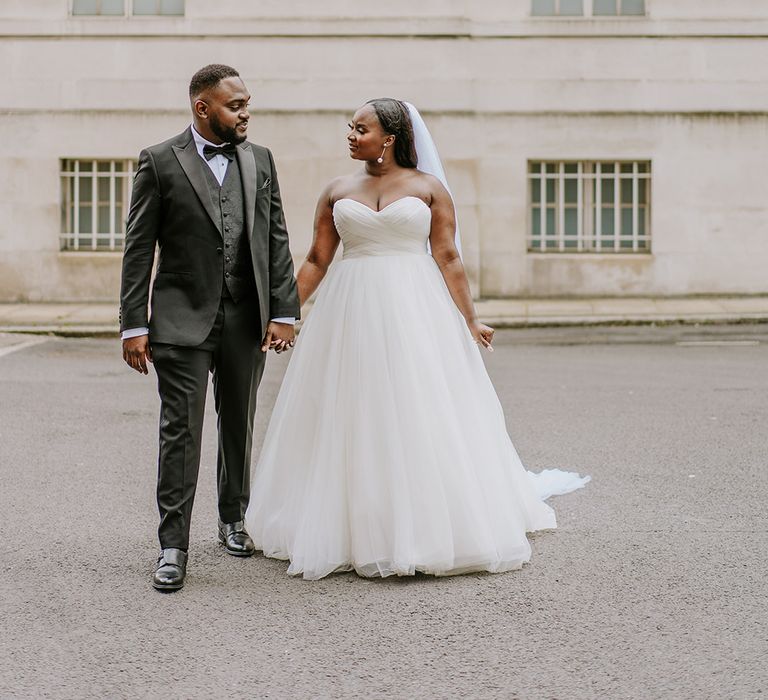 Bride & groom stand in front of white buildings after their wedding ceremony whilst holding hands