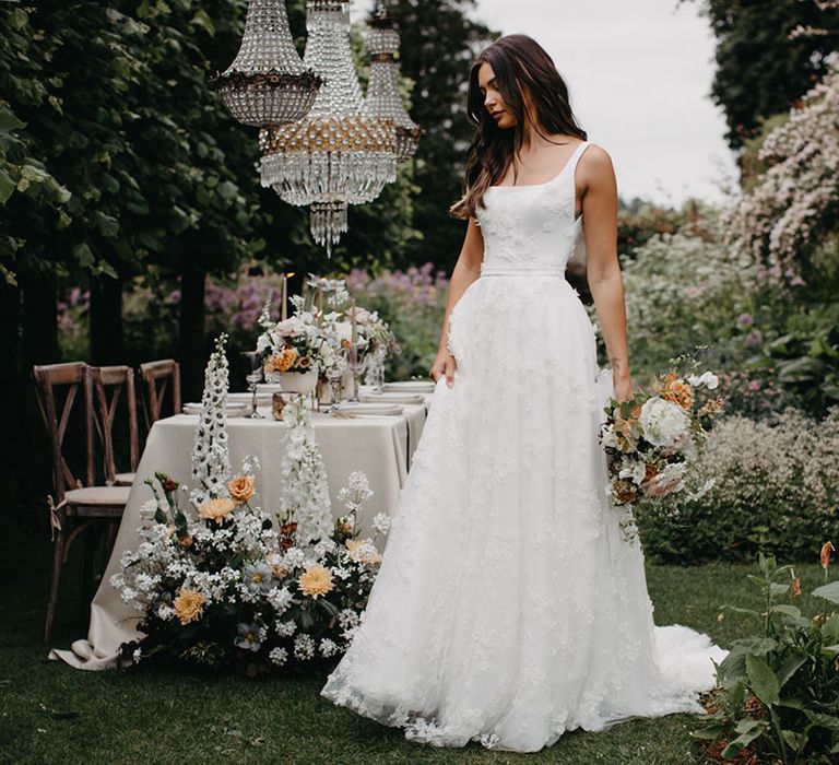 Bride in an appliqué wedding dress with fitted bodice and thin straps standing in front of an elegant outdoor wedding reception with chandelier installation