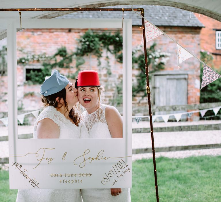 Bride kisses her bride on the cheek as they wear hats outdoors on their wedding day