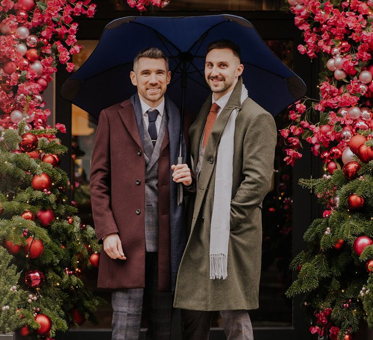 Grooms stand in front of floral archway filled with pink florals and brightly coloured baubles finished with green foliage