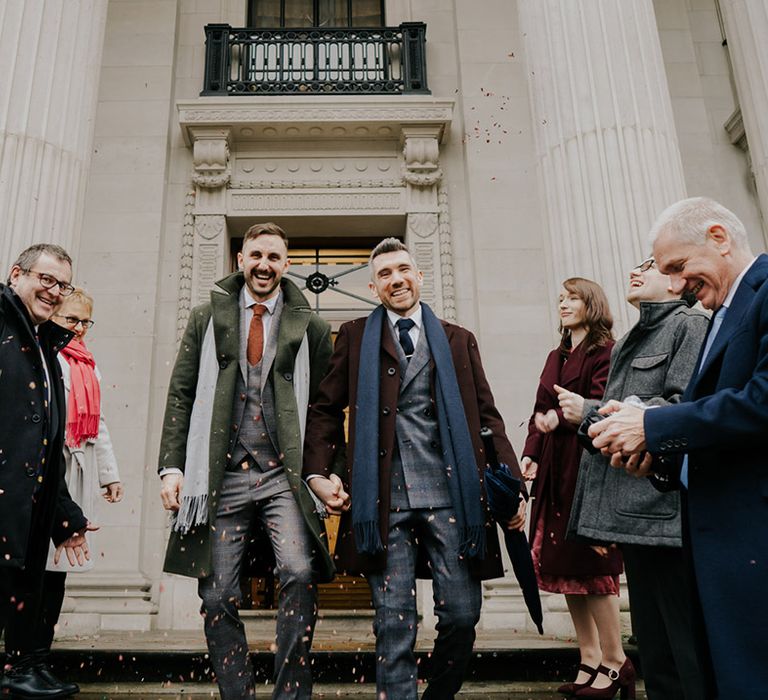 Grooms leave Old Marylebone Town Hall smiling and laughing on their wedding day as wedding guests throw confetti around them for December wedding