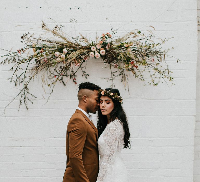 Groom in a light brown wedding suit holding hands with his bride in a lace wedding dress at the rug and flower installation altar 