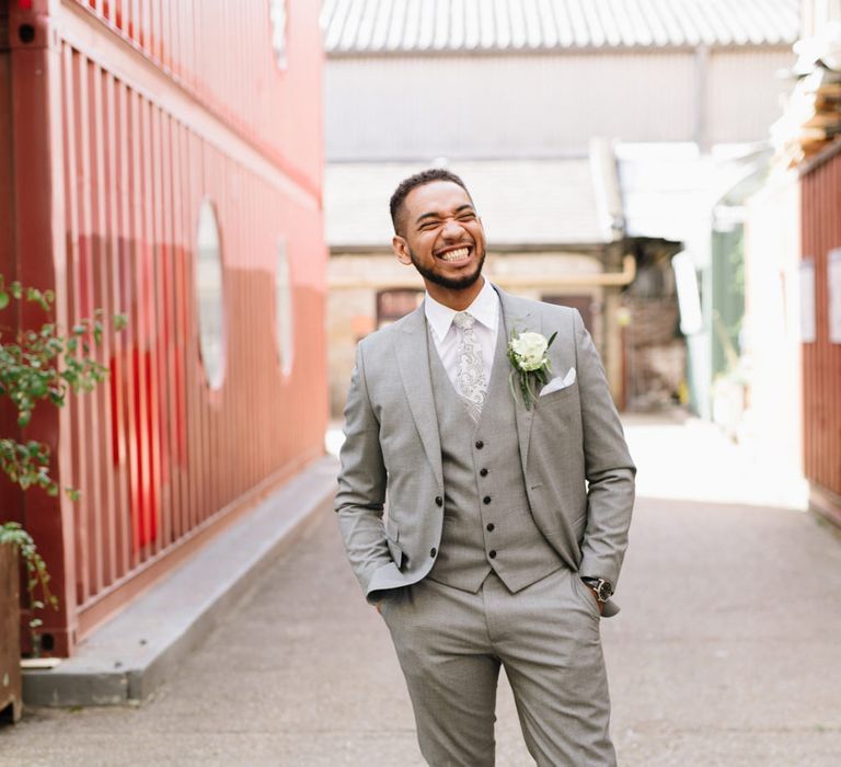 Groom in a pale grey three-piece wedding suit with paisley tie and white rose buttonhole flower 