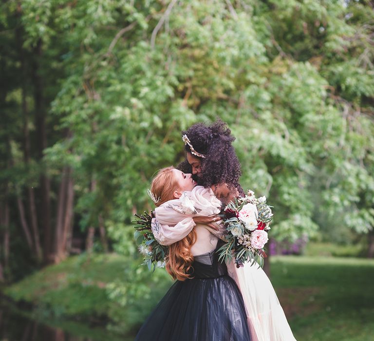 Lesbian couple embrace during wedding shoot. One bride has ginger hair and wears a black tulle dress. The other bride wears a pink tulle dress.