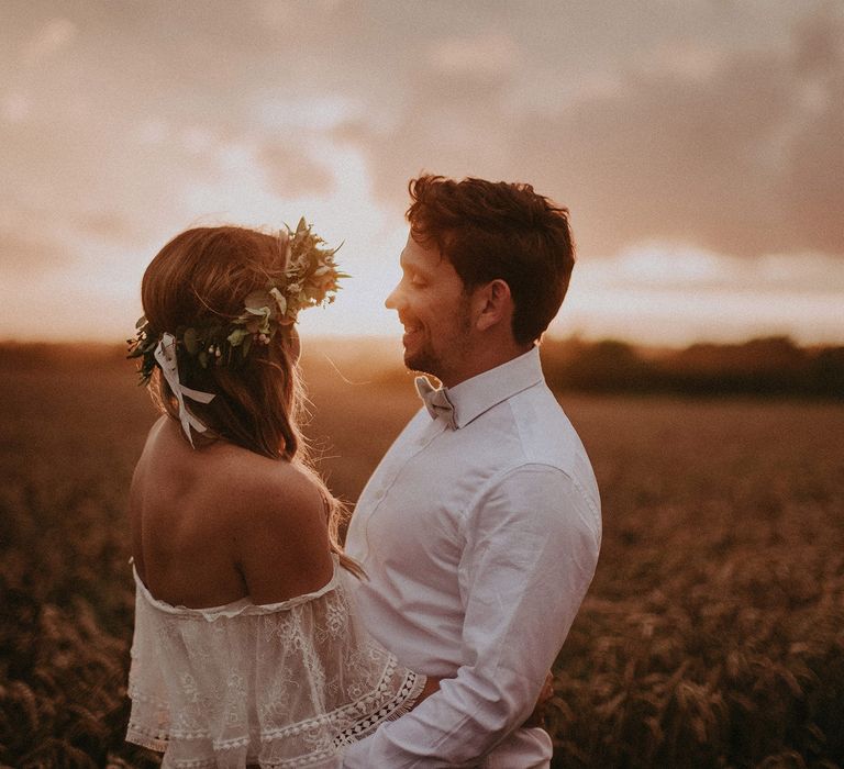 Bride in Grace Loves Lace bardot wedding dress and flower crown stands with groom in white shirt by wheat field at golden hour at Isle of Wight wedding with macrame wedding decor