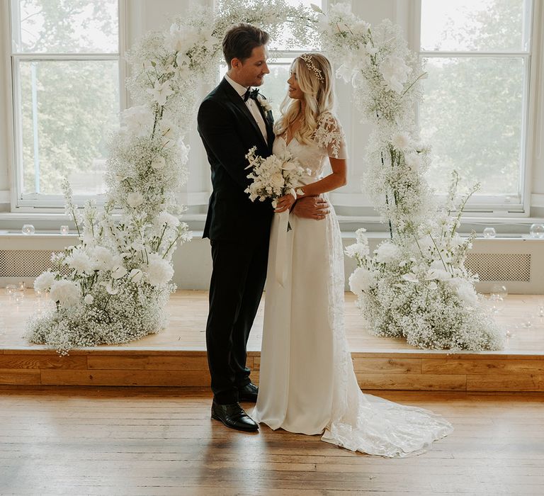 Bride and groom in black tie attire standing in front of a white flower and gypsophila arch 