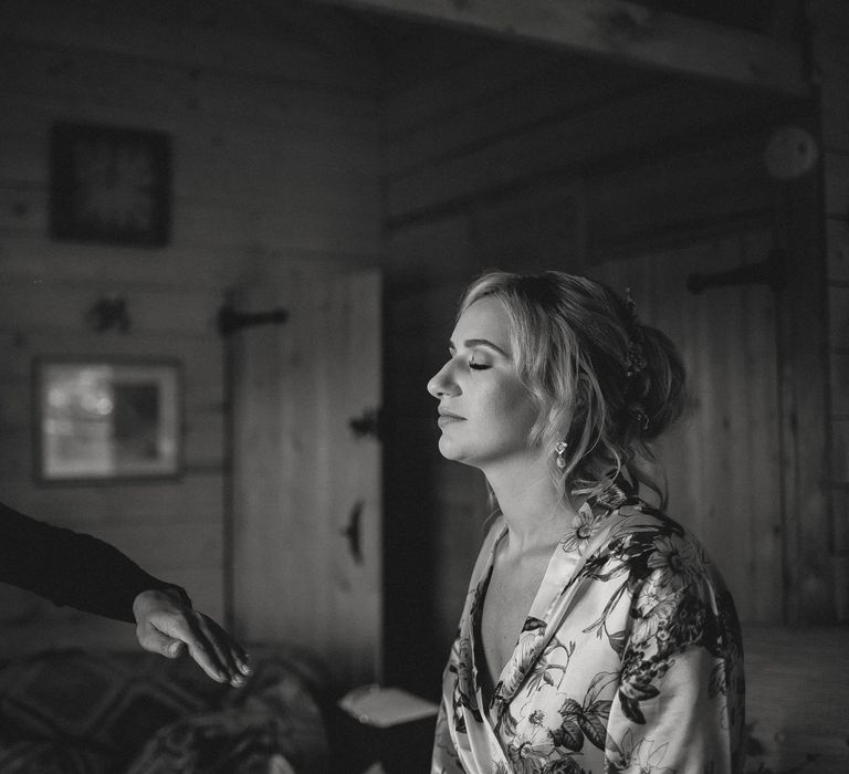 Bride in floral satin dressing gown with curled wedding updo has hair sprayed by hair stylist in bedroom before tipi wedding at Inkersall Grange Farm