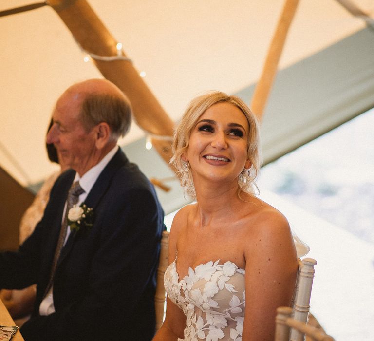 Bride in off the shoulder Enzoani wedding dress and curled wedding updo smiles up at groom as he makes speech during tipi wedding breakfast at Inkersall Grange Farm wedding