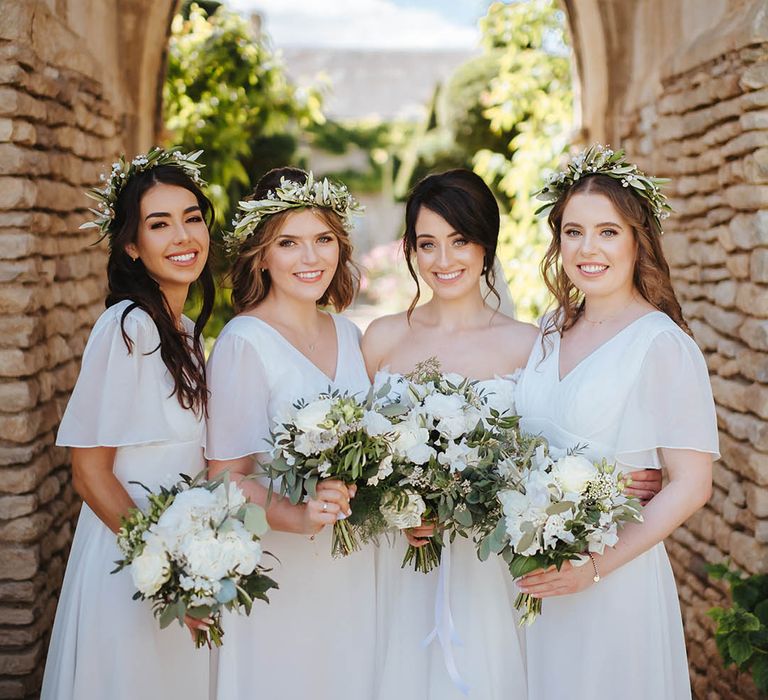 Bride stands with her bridesmaids wearing white dresses and holding white florals complete with green foliage