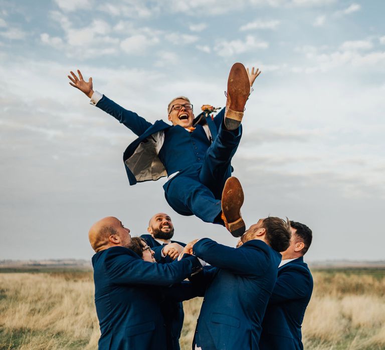 Groomsmen in Navy suits & tanned belts throw Groom in the air in open field & laugh