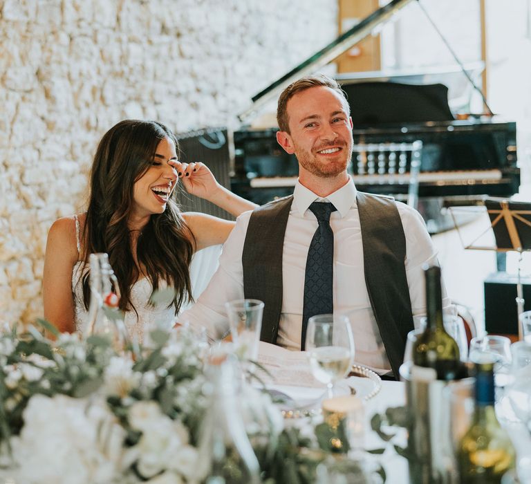 Laughing bride in Pronovias wedding dress sits holding hands with groom in white shirt and grey waistcoat during speeches at wedding reception