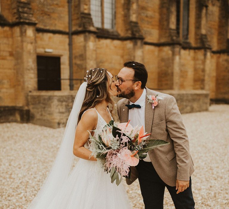 Bride & groom kiss outside church on their wedding day as bride holds floral bouquet