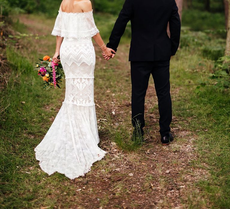 Bride in off the shoulder Grace Loves Lace wedding dress and flower crown holds colourful wedding bouquet as she looks over her shoulder whilst walking through woodland with groom in black suit