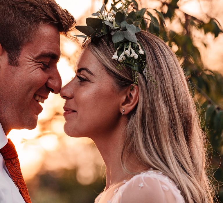 Bride in mesh sleeve wedding dress and boho bridal flower crown stands nose to nose with groom in white shirt and red tie during golden hour