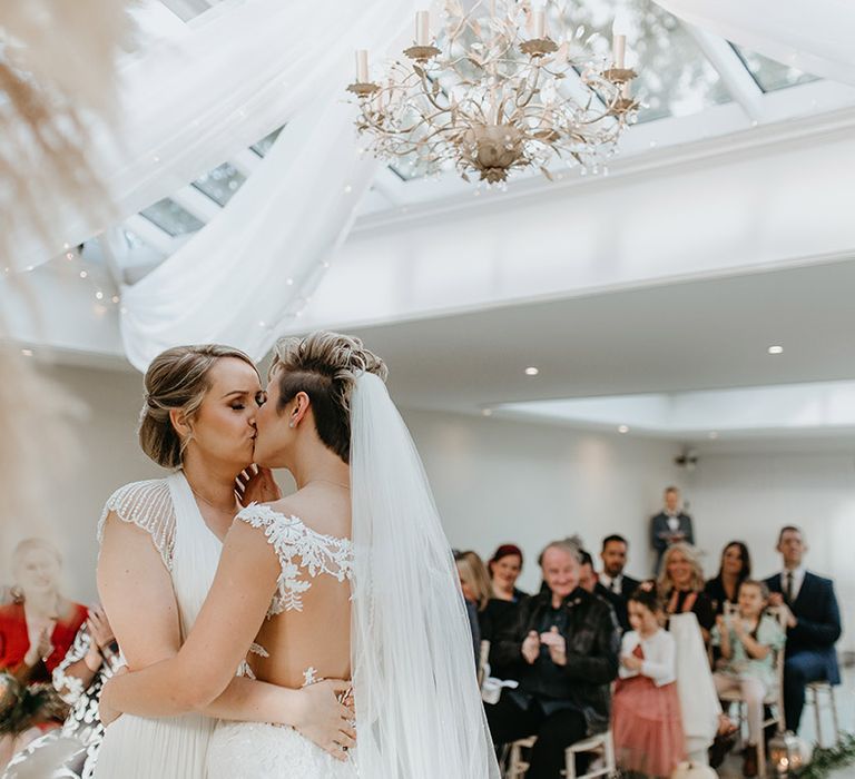 Brides kiss on their wedding day as wedding party looks on