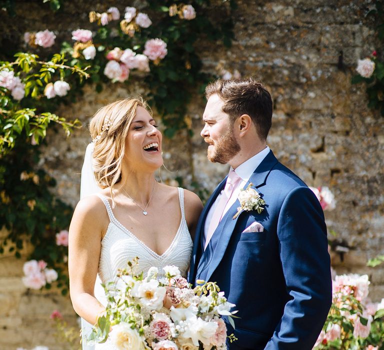 Bride in a Made With Love Bridal gown holding a white and pink bouquet laughing with her husband in a navy blue suit 