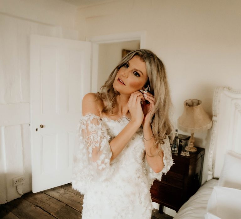 Bride in off the shoulder floral wedding dress puts in earrings whilst stood in bedroom before wedding