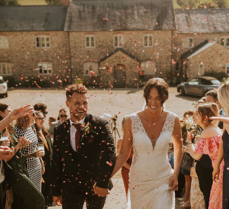 Smiling groom in dark suit and burgundy tie holds hands with bride in lace wedding dress walking up stone steps as guests throw pink confetti