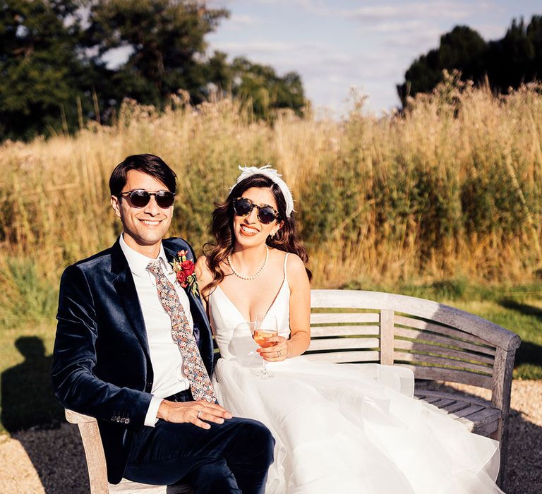 Bride in layered tulle wedding dress and feathered headband sits on wooden bench with groom in dark blue suit and floral tie as they both wear sunglasses