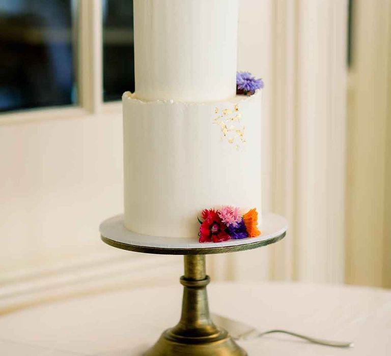 Two tier wedding cake complete with white frosting, gold leaf and bright floral decor 