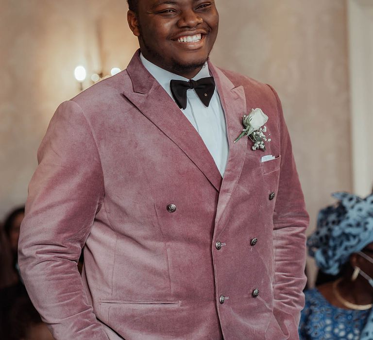 Groom wears dusky pink jacket for wedding complete with floral buttonhole and black tie 