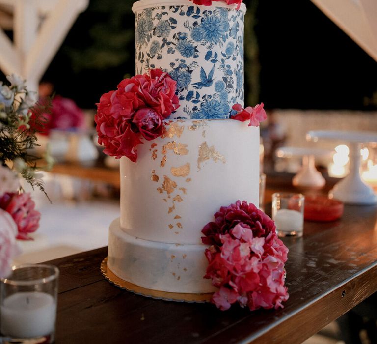 Four tier wedding cake complete with white frosting, blue floral design, gold leaf and bright red floral decor 