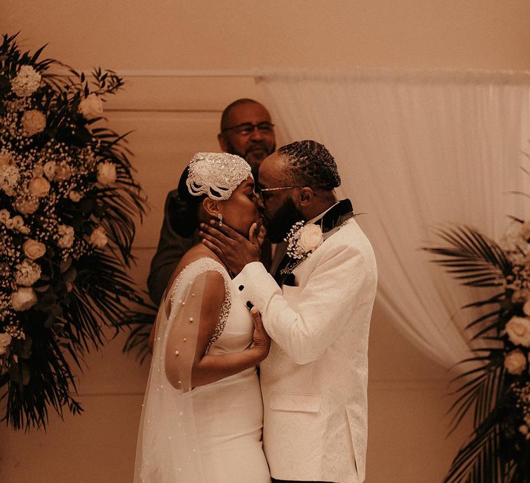 Groom in a white tuxedo jacket kissing his bride in a vintage style wedding dress with Watteau train at the altar 