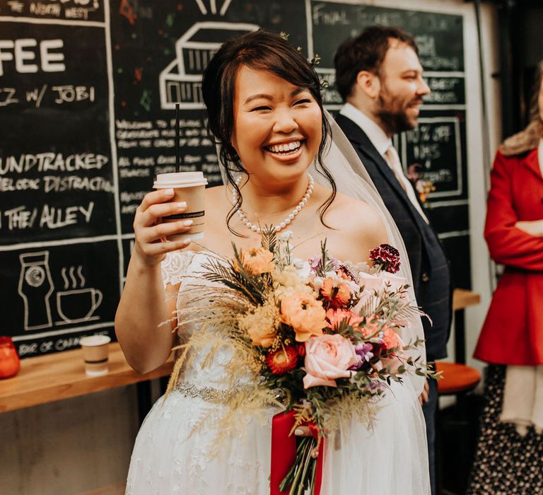 Bride in lace off the shoulder wedding dress and pearl necklace holding bright mixed orange and pink bouquet laughs whilst holding disposable coffee cup 