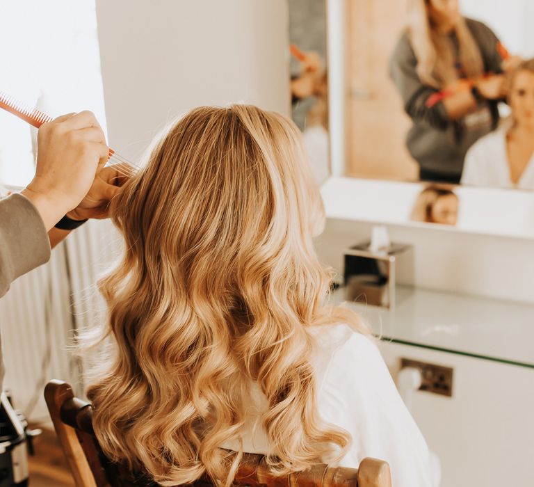 Bride with blonde curled hair sits having her hair styled before wedding