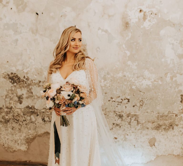 Bride in white wedding dress with pearl mesh overlay stands holding mixed pastel wedding bouquet 