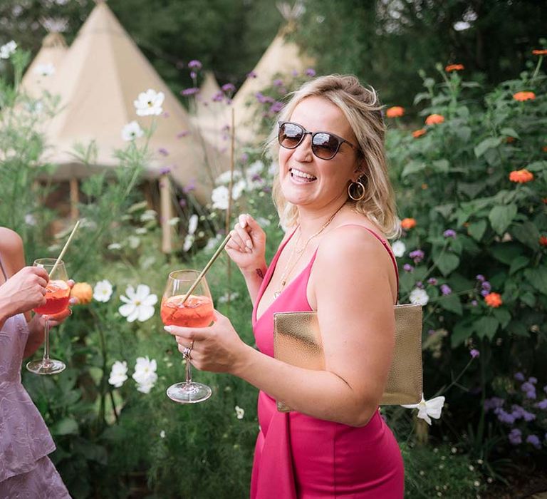 Wedding guest in lilac patterned dress and guest in bright pink dress with aperol spritz's in hand for garden wedding