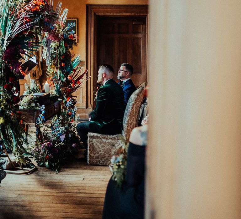 Grooms are seated for the ceremony in matching damask chairs in from of tropical floral moongate